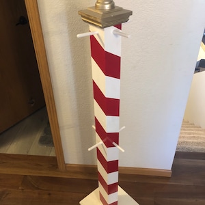 May include: A white and red striped wooden candy cane stand with a gold finial on top. The stand has four wooden arms extending out from the sides.