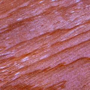May include: Close-up of a reddish-brown wood surface with a grainy texture. The surface has a slight sheen and appears to be varnished.