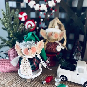 May include: Two handmade Christmas elves with red and white patterned clothing, a white truck, and red and green light bulbs on a burlap surface.