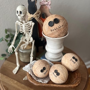 May include: A collection of Halloween decor featuring a plastic skeleton, a black wooden cat silhouette, and four round, tan, textured spheres wrapped in twine. Each sphere has two black button eyes and a stitched mouth made of dark thread.