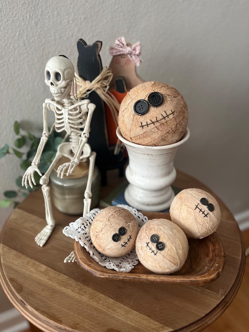 May include: A collection of Halloween decor featuring a plastic skeleton, a black wooden cat silhouette, and four round, tan, textured spheres wrapped in twine. Each sphere has two black button eyes and a stitched mouth made of dark thread.
