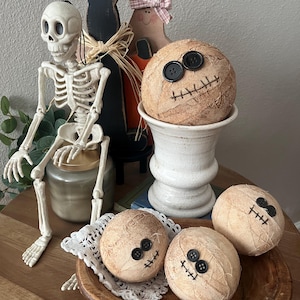 May include: Three round, beige, fabric-wrapped spheres decorated with black button eyes and stitched mouths, resembling mummies. One sphere sits in a white pedestal bowl, while two others rest in a wooden bowl with a white lace doily.