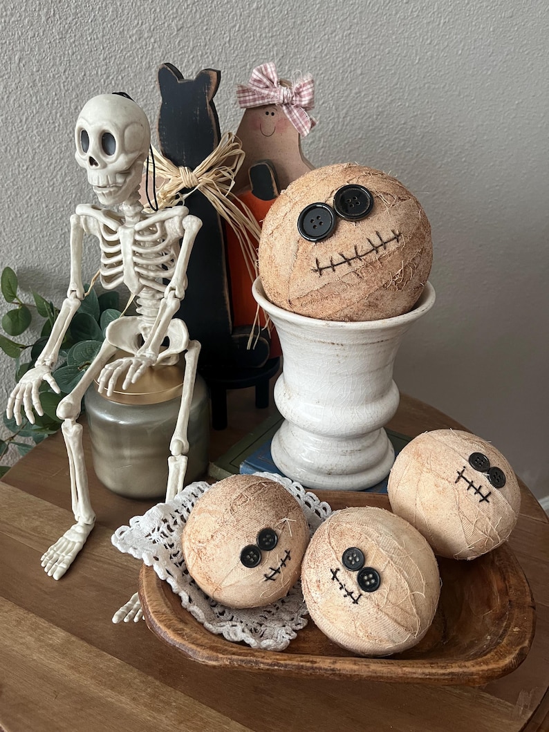 May include: Three round, beige, fabric-wrapped spheres decorated with black button eyes and stitched mouths, resembling mummies. One sphere sits in a white pedestal bowl, while two others rest in a wooden bowl with a white lace doily.