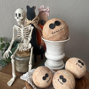 May include: A set of four round, tan, fabric-wrapped spheres featuring black button eyes and stitched mouth details. One sphere rests in a white pedestal bowl, while three others are arranged in a wooden bowl with a white lace doily.