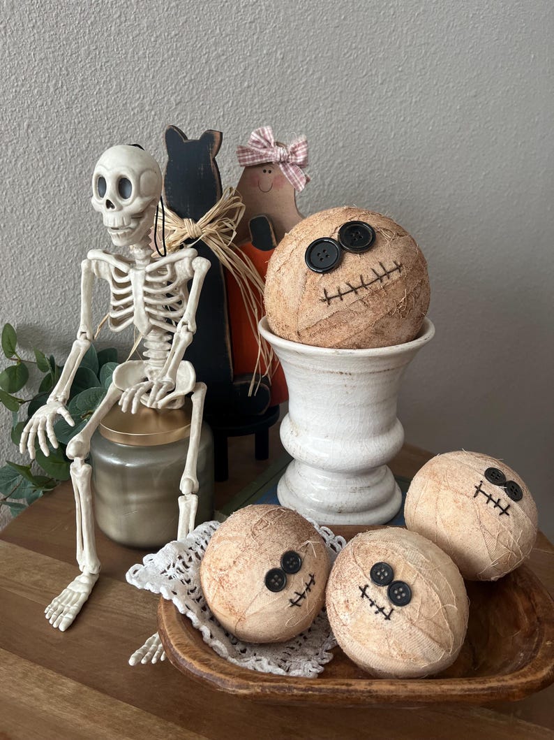 May include: A set of four round, tan, fabric-wrapped spheres featuring black button eyes and stitched mouth details. One sphere rests in a white pedestal bowl, while three others are arranged in a wooden bowl with a white lace doily.