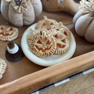 May include: A white plate with a variety of pumpkin-shaped pie pops with orange filling and a cinnamon-dusted crust. The plate is on a wooden tray with a black and white checkered cloth.