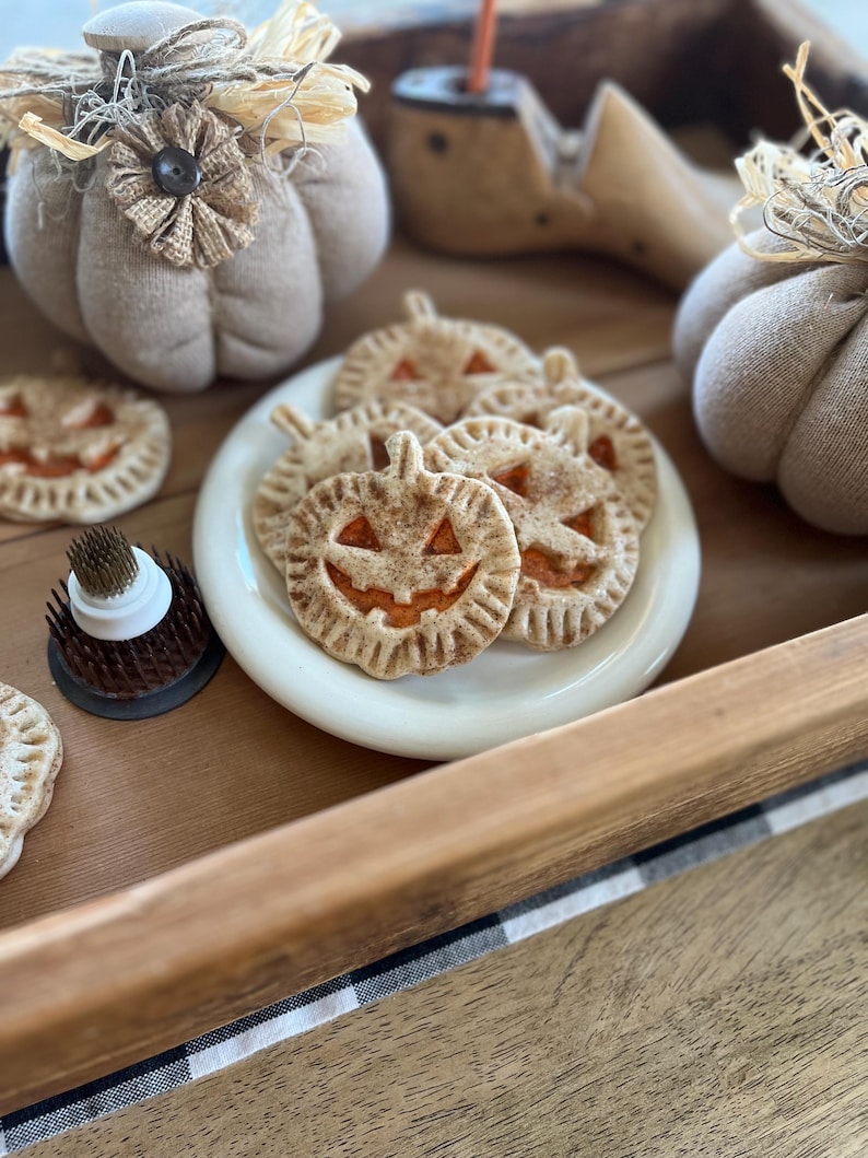 May include: A white plate with a variety of pumpkin-shaped pie pops with orange filling and a cinnamon-dusted crust. The plate is on a wooden tray with a black and white checkered cloth.