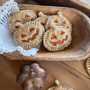 May include: A wooden bowl filled with pumpkin-shaped cookies decorated with orange icing and a jack-o'-lantern face. The cookies are on a white doily and there is a small brown pumpkin-shaped mold next to the bowl.