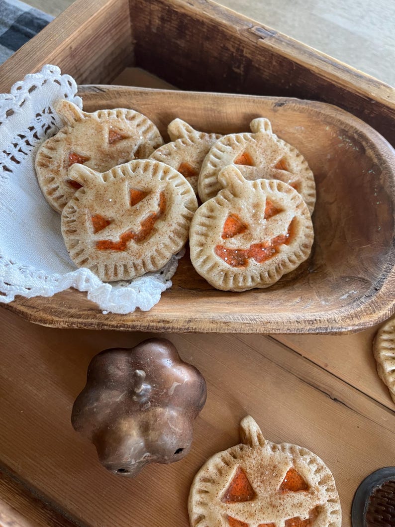 May include: A wooden bowl filled with pumpkin-shaped cookies decorated with orange icing and a jack-o'-lantern face. The cookies are on a white doily and there is a small brown pumpkin-shaped mold next to the bowl.