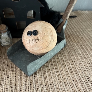 May include: A round, tan, fabric-wrapped ball with two black button eyes and a stitched mouth, resting in a galvanized metal scoop. Beside it sits a small glass bottle with a cork stopper labeled Ghost's Breath and a black wooden house cutout.