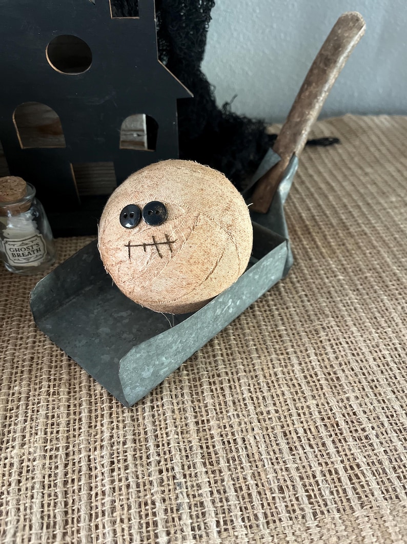 May include: A round, tan, fabric-wrapped ball with two black button eyes and a stitched mouth, resting in a galvanized metal scoop. Beside it sits a small glass bottle with a cork stopper labeled Ghost's Breath and a black wooden house cutout.