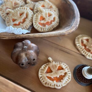 May include: A wooden bowl filled with pumpkin-shaped pie crusts with orange filling and a jack-o'-lantern face. There is a small ceramic pumpkin and another pie crust on a wooden surface.