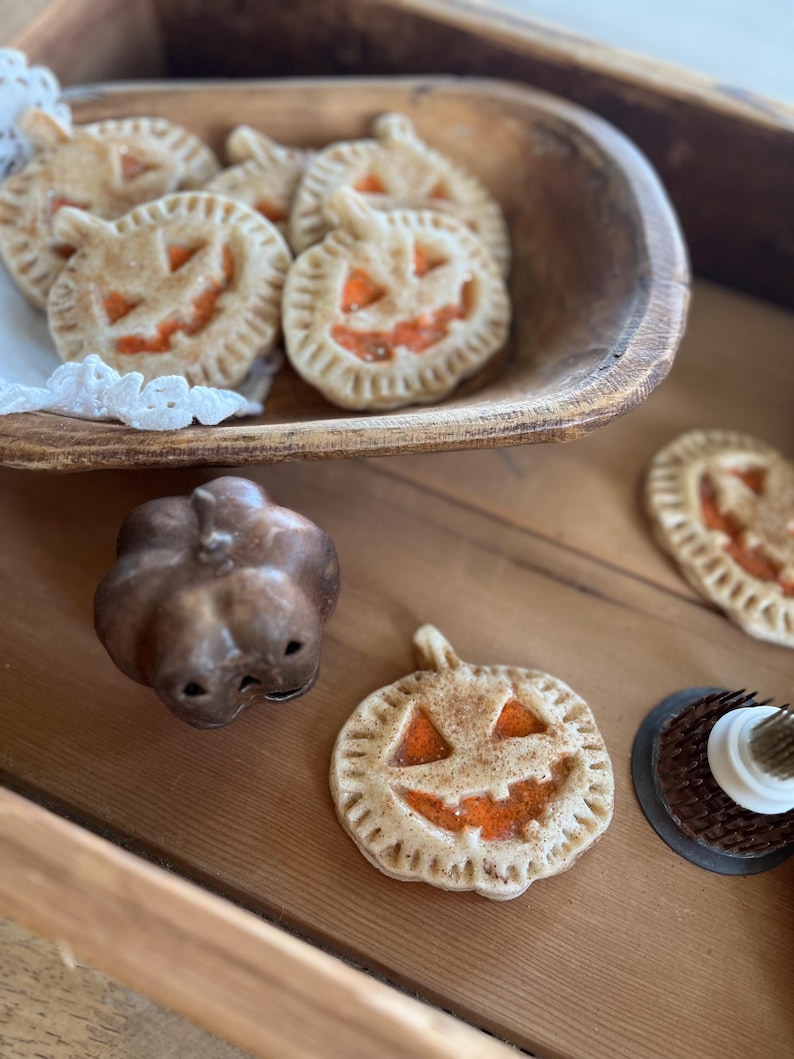 May include: A wooden bowl filled with pumpkin-shaped pie crusts with orange filling and a jack-o'-lantern face. There is a small ceramic pumpkin and another pie crust on a wooden surface.