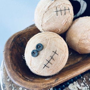May include: Three spherical fabric-wrapped decorations in a light beige color. Each sphere features two dark gray buttons attached as eyes and black stitched lines resembling a mouth. The fabric has a frayed, textured appearance.