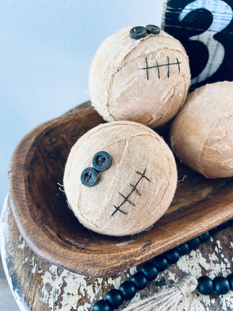 May include: Three spherical fabric-wrapped decorations in a light beige color. Each sphere features two dark gray buttons attached as eyes and black stitched lines resembling a mouth. The fabric has a frayed, textured appearance.