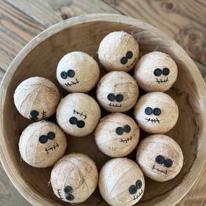 May include: A collection of eleven spherical objects wrapped in beige fabric strips to resemble mummies. Each sphere features two black button eyes and a stitched black thread mouth, arranged together inside a large, round, light brown wooden bowl.