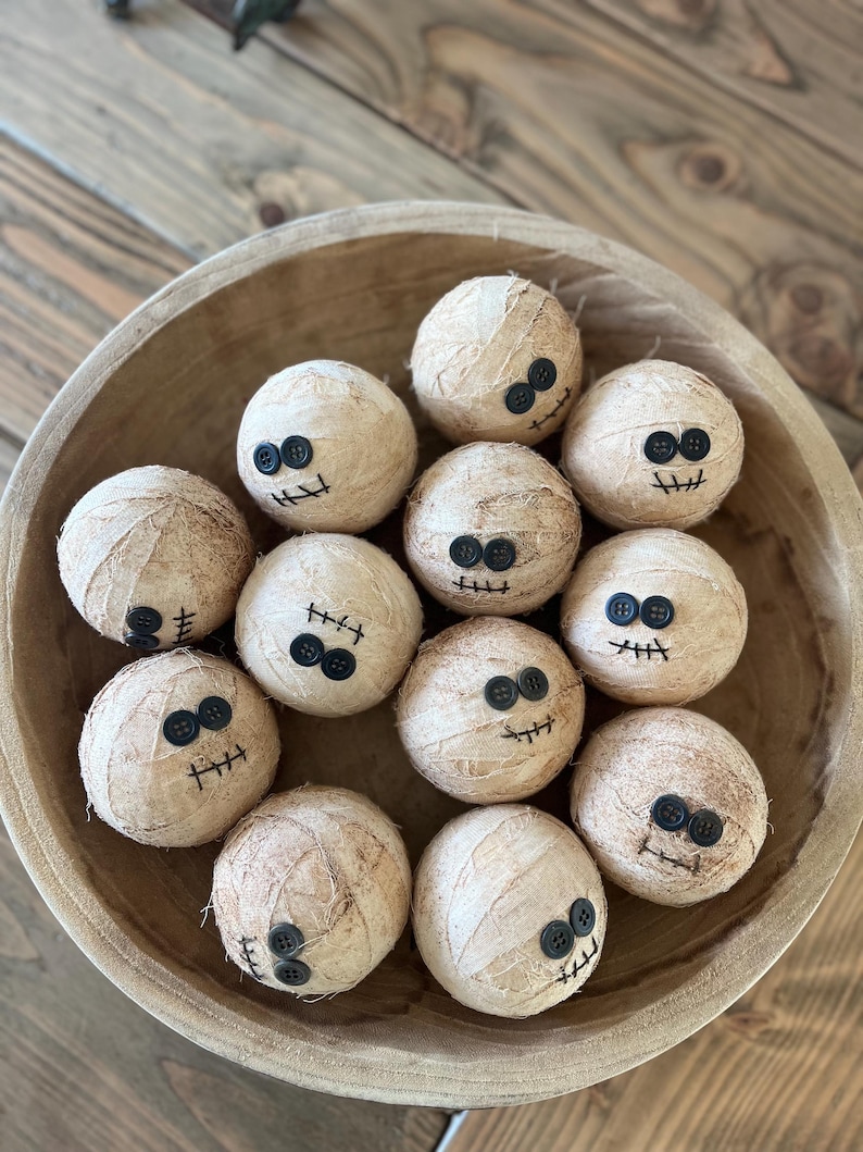May include: A collection of eleven spherical objects wrapped in beige fabric strips to resemble mummies. Each sphere features two black button eyes and a stitched black thread mouth, arranged together inside a large, round, light brown wooden bowl.