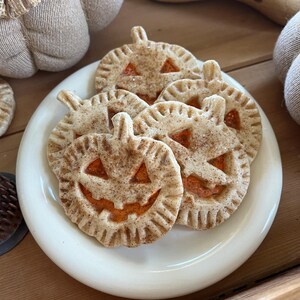 May include: A white plate with six pumpkin-shaped pie crusts with orange filling and a cinnamon-sugar topping. The pie crusts are shaped like jack-o'-lanterns with a smiling face.