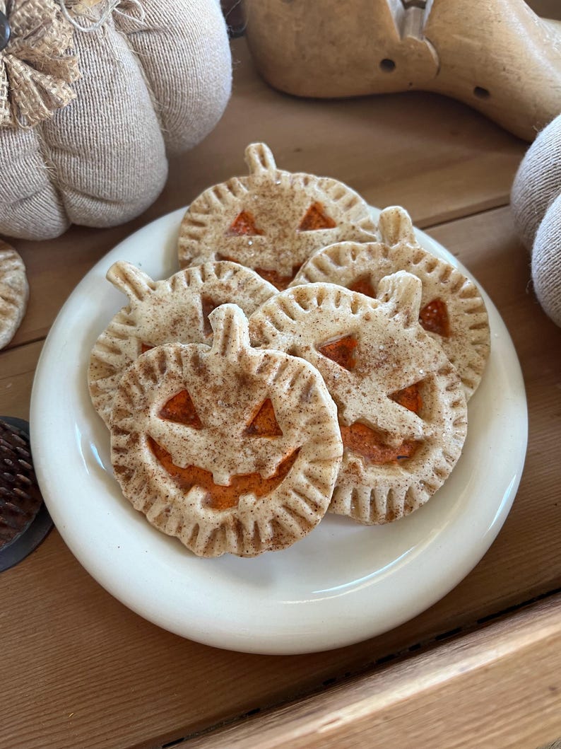 May include: A white plate with six pumpkin-shaped pie crusts with orange filling and a cinnamon-sugar topping. The pie crusts are shaped like jack-o'-lanterns with a smiling face.