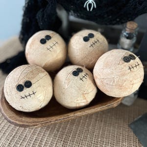 May include: Five spherical objects wrapped in beige fabric to resemble mummies, each featuring two small black button eyes and a stitched black mouth. These round decorations are arranged together in a small, shallow, dark brown wooden bowl.