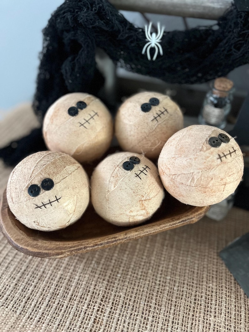 May include: Five spherical objects wrapped in beige fabric to resemble mummies, each featuring two small black button eyes and a stitched black mouth. These round decorations are arranged together in a small, shallow, dark brown wooden bowl.