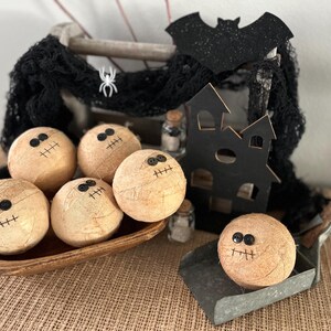 May include: Five round, tan, textured spheres decorated with black button eyes and stitched mouths, resembling mummies. These are arranged in a wooden bowl and a metal scoop, accompanied by a black wooden house cutout and black netting.