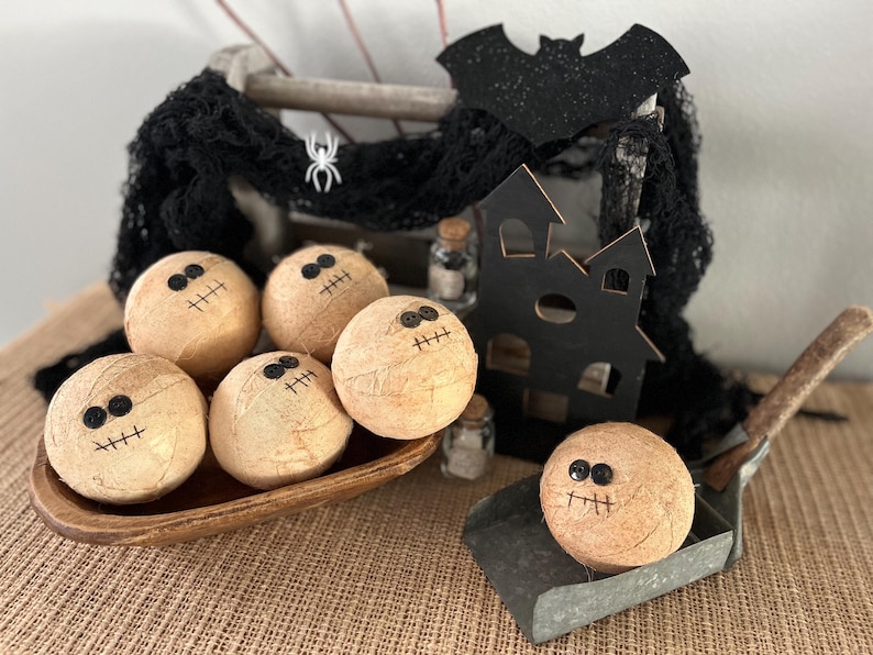 May include: Five round, tan, textured spheres decorated with black button eyes and stitched mouths, resembling mummies. These are arranged in a wooden bowl and a metal scoop, accompanied by a black wooden house cutout and black netting.