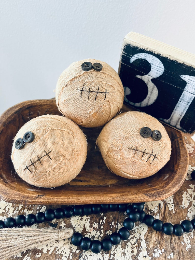 May include: Three spherical fabric-wrapped decorations in a tan color, each featuring two dark button eyes and a stitched mouth. The items are arranged in a brown wooden bowl next to a black and white wooden block displaying the number 31.