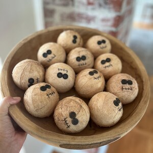 May include: A large, round, light brown wooden bowl filled with twelve spherical, tan-colored fabric-wrapped balls. Each ball features two black button eyes and a stitched black thread mouth, creating a simple, uniform facial expression on every item.