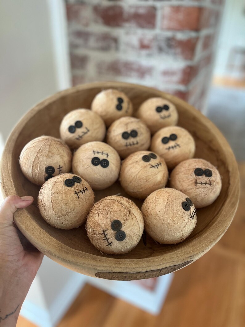 May include: A large, round, light brown wooden bowl filled with twelve spherical, tan-colored fabric-wrapped balls. Each ball features two black button eyes and a stitched black thread mouth, creating a simple, uniform facial expression on every item.