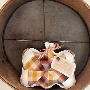 May include: A fabric pumpkin decoration featuring a patchwork design of white, orange, and brown plaid fabric. The pumpkin is topped with a thin orange and white striped ribbon and a small metal bell attached to a rectangular fabric tag.