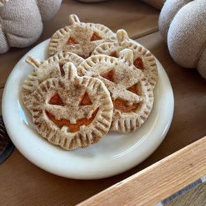 May include: A white plate with five pumpkin-shaped cookies with orange icing and a jack-o'-lantern face. The cookies are on a wooden surface with a burlap-covered pumpkin in the background.