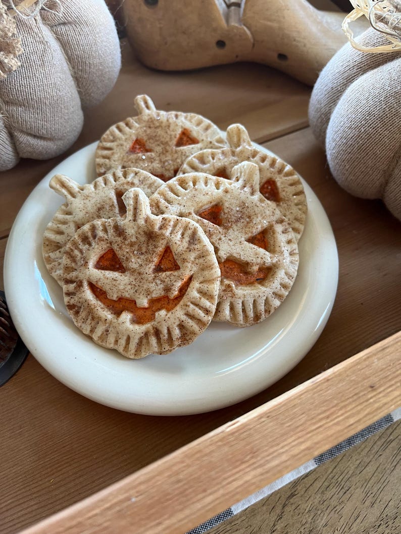 May include: A white plate with five pumpkin-shaped cookies with orange icing and a jack-o'-lantern face. The cookies are on a wooden surface with a burlap-covered pumpkin in the background.