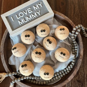 May include: Eight round, tan, fabric-wrapped spheres resembling mummies, each featuring two black button eyes and a stitched mouth. A light gray felt letter board displays the white text "I LOVE MY MUMMY" above the spheres on a wooden tray.