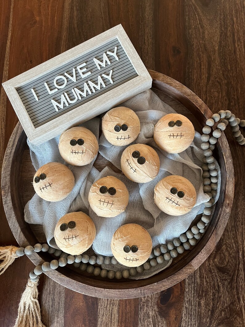 May include: Eight round, tan, fabric-wrapped spheres resembling mummies, each featuring two black button eyes and a stitched mouth. A light gray felt letter board displays the white text "I LOVE MY MUMMY" above the spheres on a wooden tray.