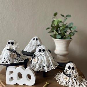 May include: Four white lace ghost figures with black felt eyes, each wearing a small black ribbon and a round tag printed with the text Trick or Treat. A white wooden block with the word BOO and a small yellow bone-shaped object are included.