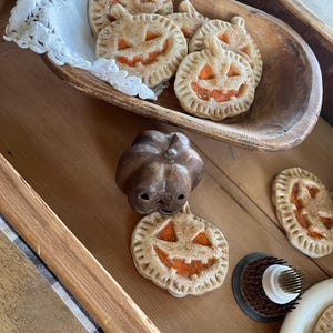 May include: A wooden tray with a variety of pumpkin-shaped cookies. The cookies are decorated with orange icing and have a smiling face. There is also a small pumpkin-shaped cookie cutter on the tray.