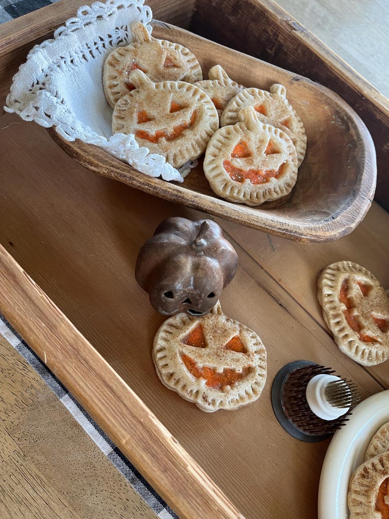 May include: A wooden tray with a variety of pumpkin-shaped cookies. The cookies are decorated with orange icing and have a smiling face. There is also a small pumpkin-shaped cookie cutter on the tray.