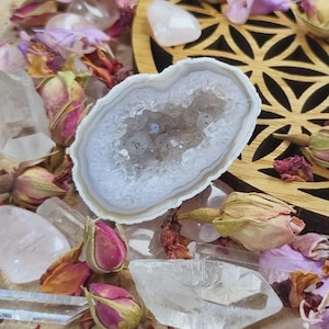 Könnte beinhalten: Ein weiß-grauer Geodenkristall mit funkelndem Inneren, umgeben von Bergkristallen, Rosenquarz, getrockneten Rosenknospen und Blütenblättern. Die Geode liegt auf einer Holzoberfläche mit floralem Design.