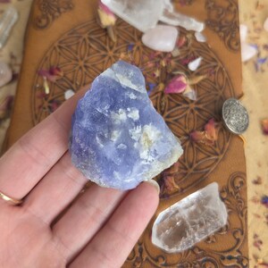 May include: A raw, translucent blue chalcedony crystal with irregular, jagged edges and white mineral inclusions. The stone features a multifaceted, crystalline surface texture with varying shades of light to deep periwinkle blue throughout.