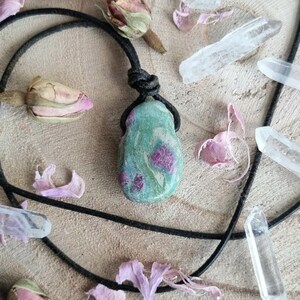 Peut inclure: Un collier pendentif artisanal avec une pierre verte et rose, suspendue à un cordon noir. La pierre a un aspect marbré avec des inclusions roses. Le collier est présenté avec des fleurs séchées et des cristaux de quartz clair.