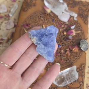 May include: A raw, triangular-shaped blue chalcedony crystal with a translucent, textured surface and jagged edges. The stone displays varying shades of periwinkle and light blue, featuring natural crystalline formations and a rough, unpolished finish.