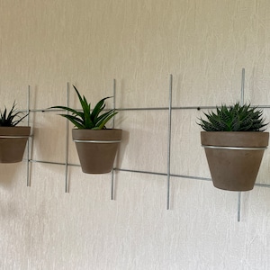 Pode incluir: Quatro vasos marrons com suculentas verdes pendurados em uma grade de metal na parede. Cada vaso tem uma borda prateada. A grade é montada em uma parede texturizada esbranquiçada.