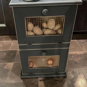 May include: A black wooden storage cabinet with two compartments. The top compartment contains several potatoes, and the bottom compartment holds two onions. Each compartment has a wire mesh door and a round knob.