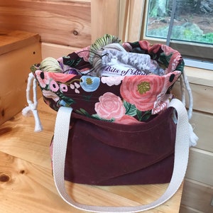 May include: A burgundy canvas tote bag with a floral print lining and white straps. The bag is filled with yarn and has a label that reads "Bits of Boo"