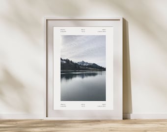 May include: A framed photograph of a mountain lake with a reflection of the sky and clouds in the water. The mountains are covered in trees and snow. The photo is in a white frame with a white mount.