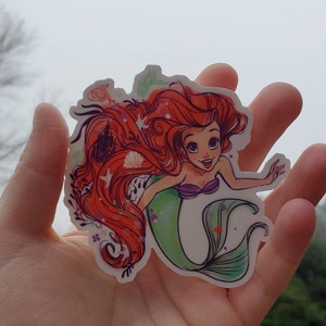 Op de afbeelding: Een sticker met een cartoonillustratie van Ariel, de Kleine Zeemeermin, met haar lange rode haar, haar groene staart en haar paarse schelpbeha. De sticker is omgeven door een witte rand en heeft een verscheidenheid aan kleine details, waaronder schelpen, zeesterren en bloemen.