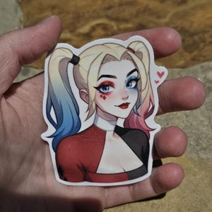 May include: A sticker featuring a cartoon illustration of Harley Quinn. She has blue and pink hair, red and black clothing, and a mischievous expression. She has red lipstick and blue eyeshadow. There are two small red hearts in the upper right corner of the sticker.