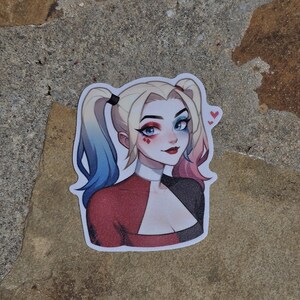 May include: A sticker featuring a cartoon illustration of Harley Quinn. She has blue and pink hair, red and black clothing, and a mischievous expression. She is wearing red lipstick and has hearts drawn on her cheeks.