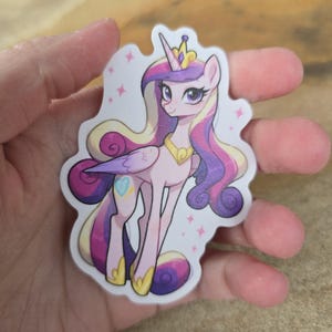 May include: A sticker featuring a cartoon pony with a crown, purple and pink hair, and a heart-shaped cutie mark. The pony has a gold necklace and is set against a white background with pink star accents. The sticker is held in a hand.